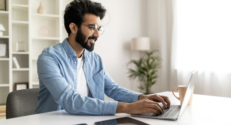 Smiling Indian Male Freelancer Working With Laptop At Home Office